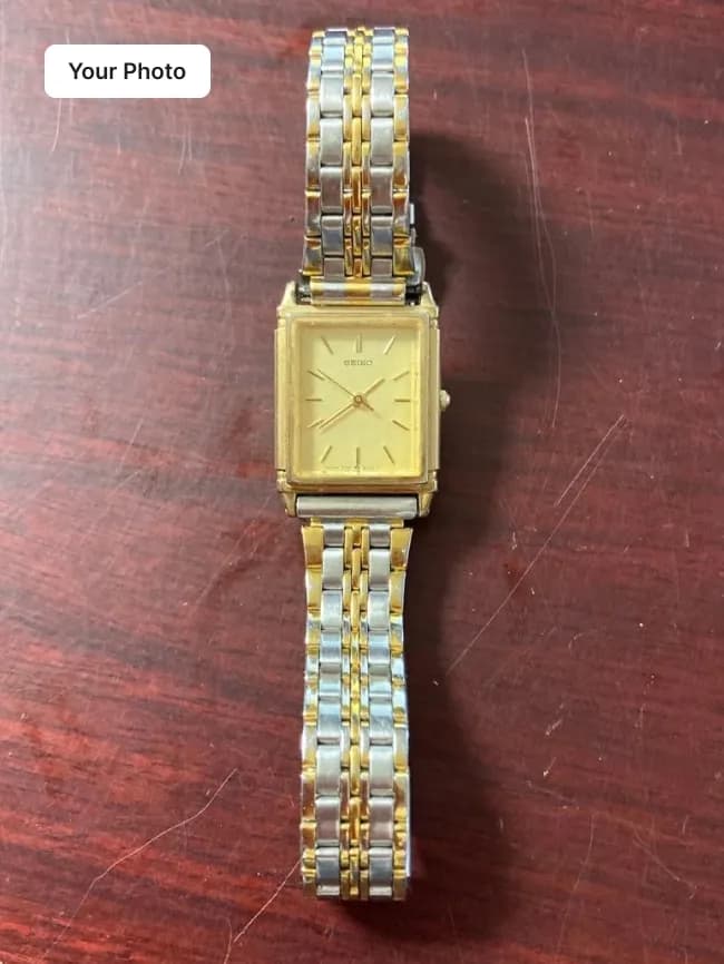 Plain phone photo of a gold and steel rectangular watch on a dark wooden table